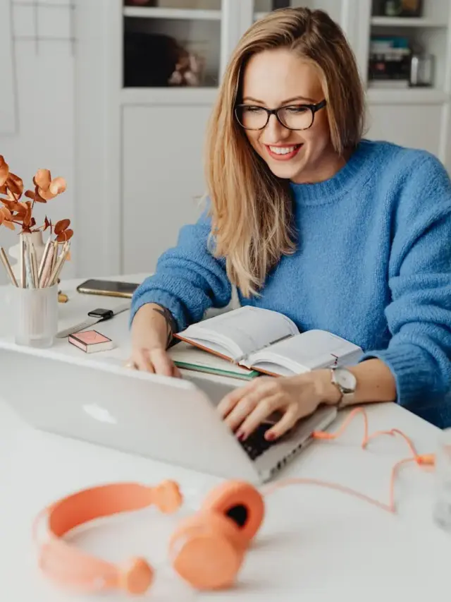 A cheerful woman in a blue sweater working remotely with a laptop in a cozy home setting.